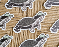 A close-up flat lay of several die-cut stickers featuring illustrated turtles wearing light green hats, placed on a woven tan rope surface. Linocut Design