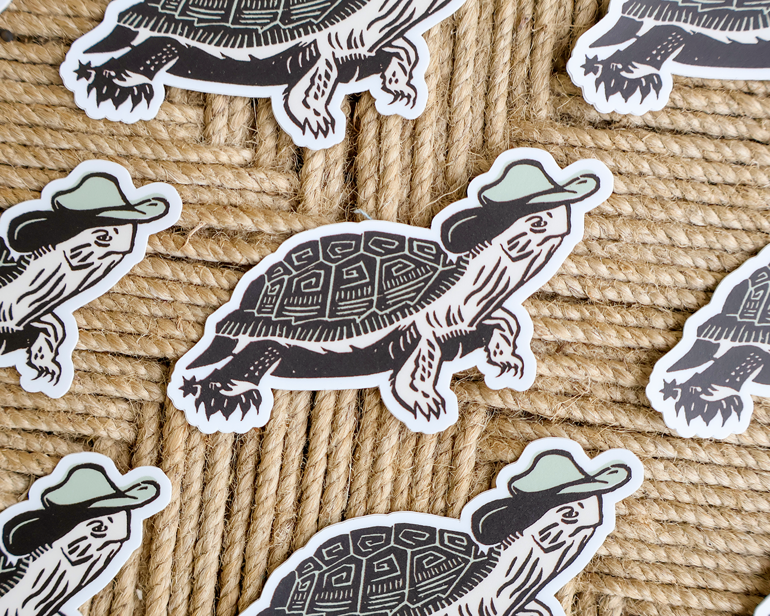 A close-up flat lay of several die-cut stickers featuring illustrated turtles wearing light green hats, placed on a woven tan rope surface. Linocut Design
