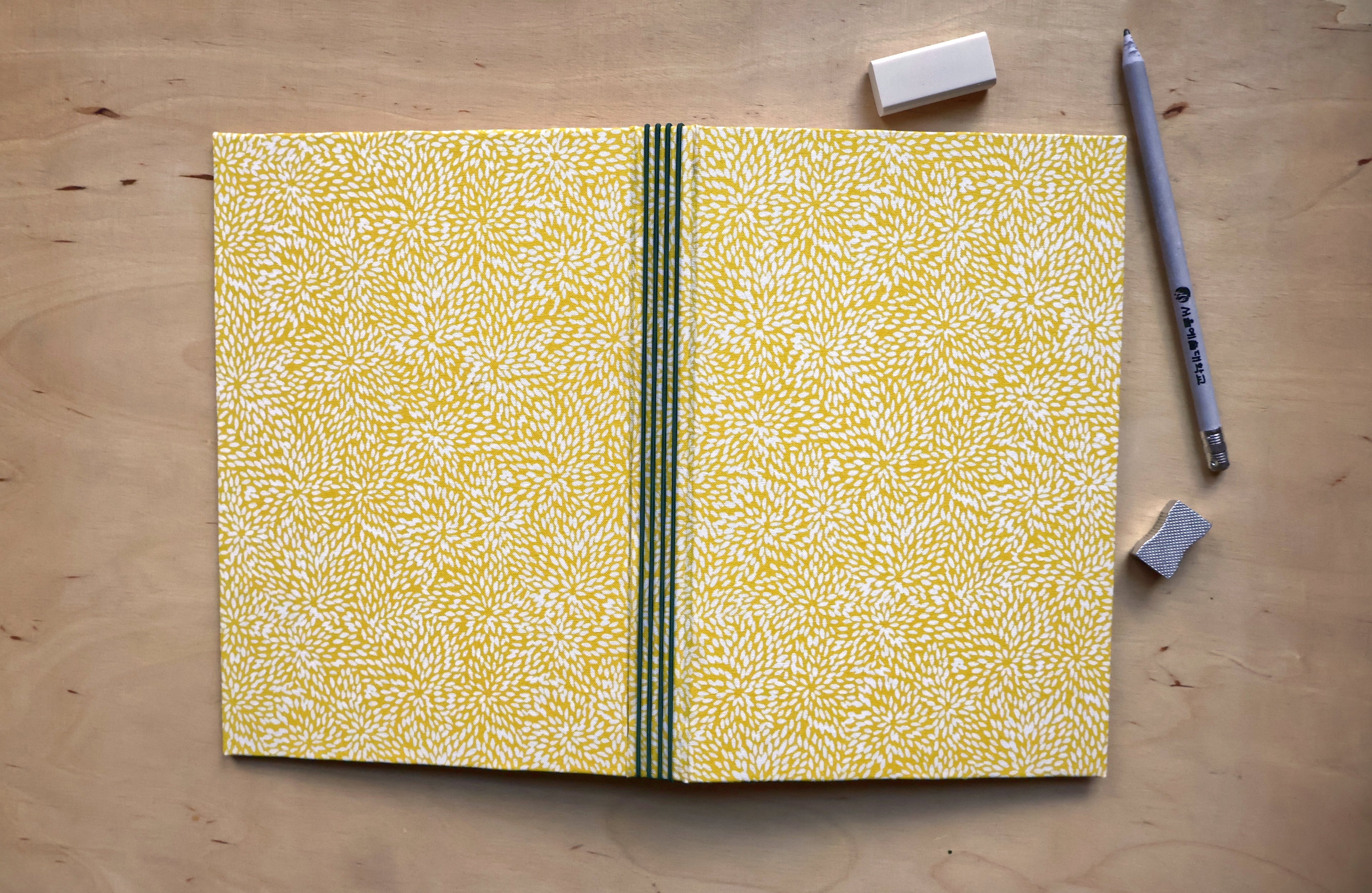A yellow and white patterned notebook with a green and black center stripe, laid flat on a light wood desk with a pencil, erasers, and a small square object nearby.