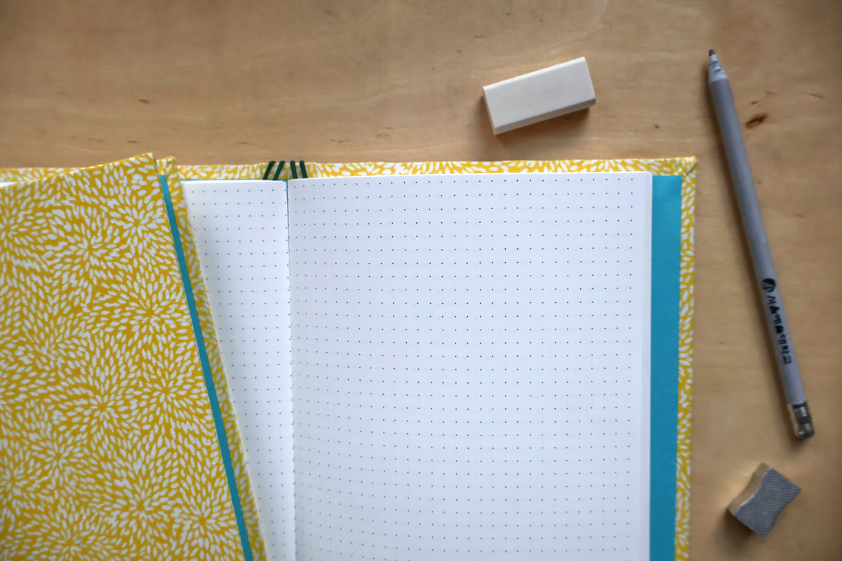 An open bullet journal with white dotted pages, a yellow and white patterned cover, and a turquoise spine, styled on a wooden desk with a pencil, eraser, and rubber.