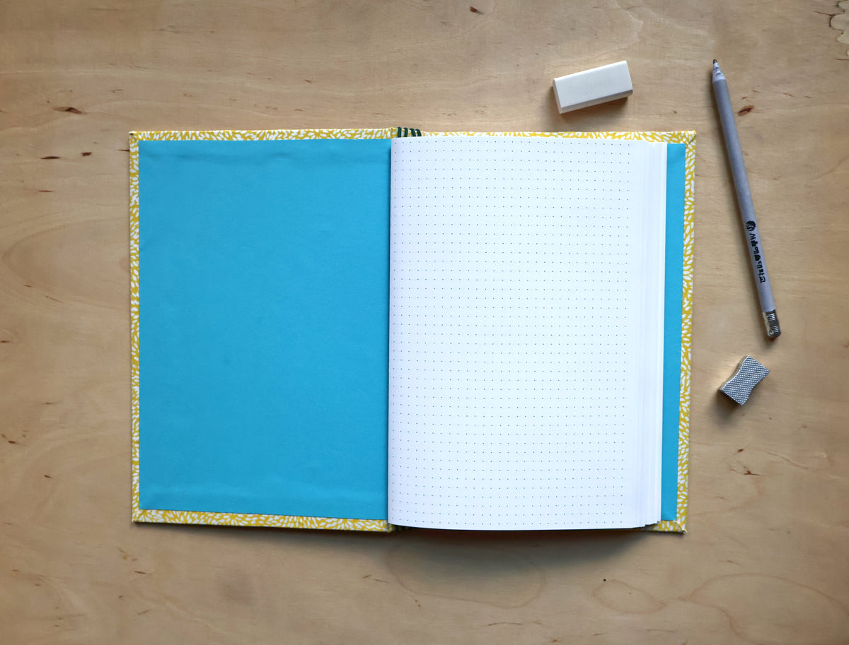 An open notebook with a bright blue left page, white grid right page, and a gold patterned cover border, placed on a light wood desk with a pencil, erasers, and a small square accessory nearby.