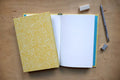 A yellow and white patterned notebook with an open grid page, styled on a wooden desk with a pencil, erasers, and a small accessory.