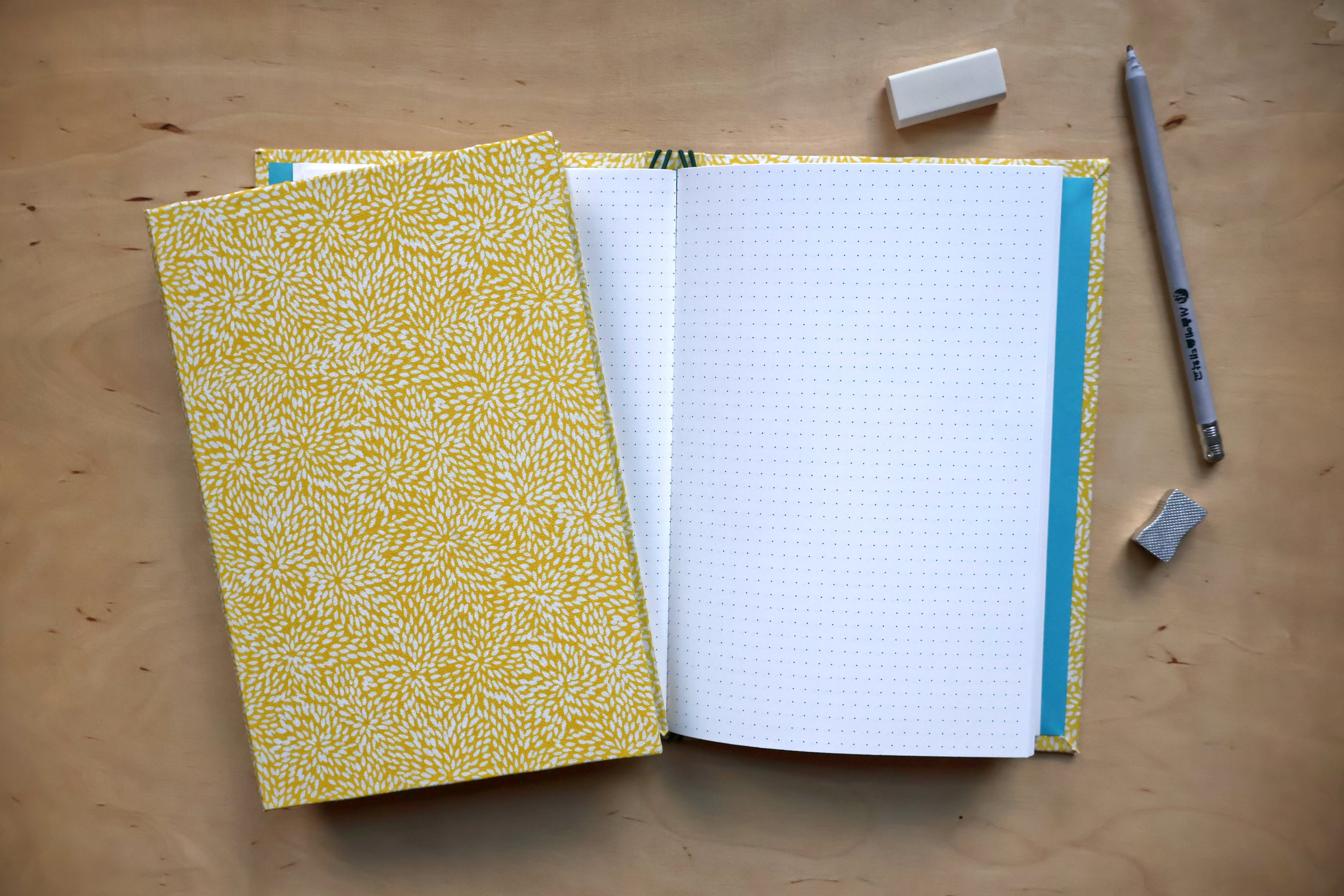 A yellow and white patterned notebook with an open grid page, styled on a wooden desk with a pencil, erasers, and a small accessory.