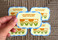 A hand holds a round sticker or coaster with a retro design, featuring orange tulips and the text 'YOU DECIDE WHAT GETS TO BLOOM' on a cream background with a blue border. Four identical items are arranged on a textured brown woven surface.