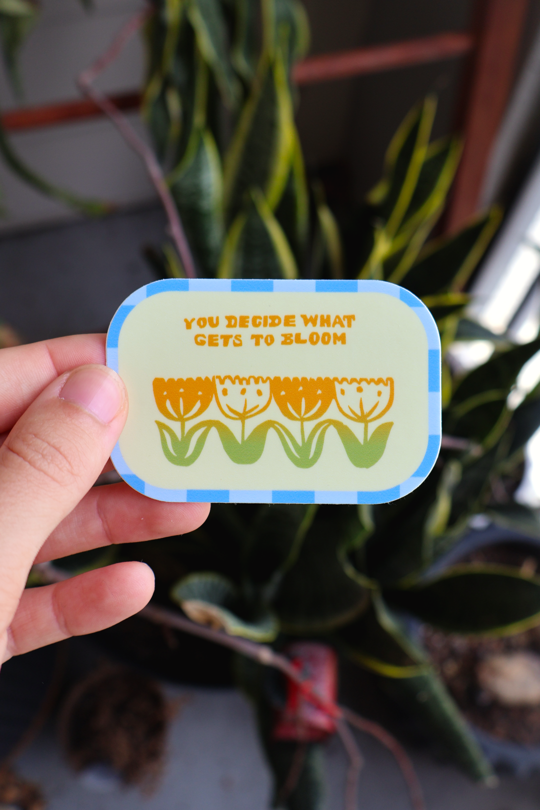 A hand holds a small rounded rectangular sticker with a blue border and a yellow and green floral design. The sticker reads, 'YOU DECIDE WHAT GETS TO BLOOM.