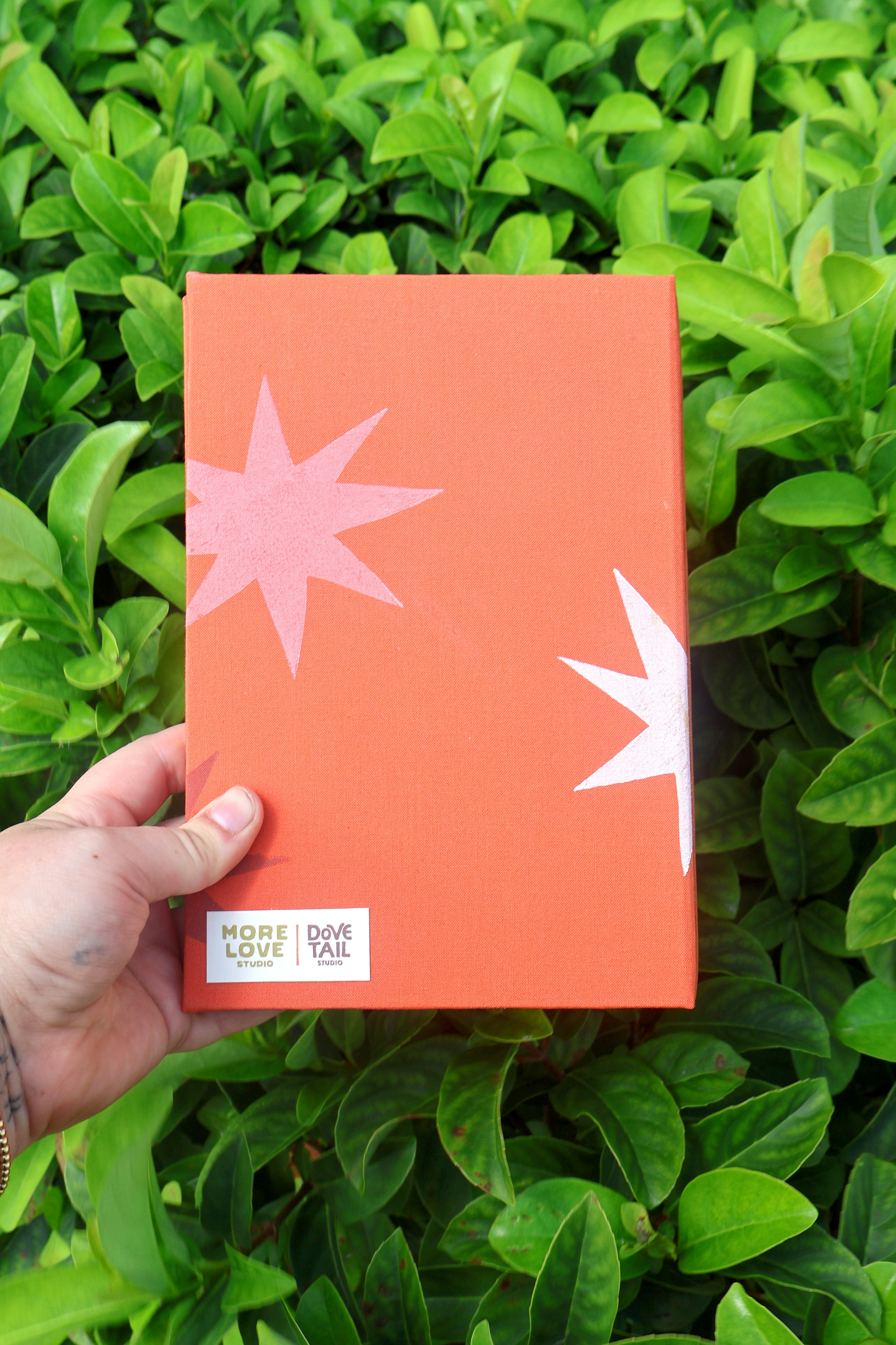 A hand holds a coral-orange book or notebook against a background of green leaves. The cover features a pink starburst on the left and a white starburst on the right, with a white label at the bottom left that reads 'MORE LOVE STUDIO and  DOVE TAIL STUDIO'. hand bound and linocut printed