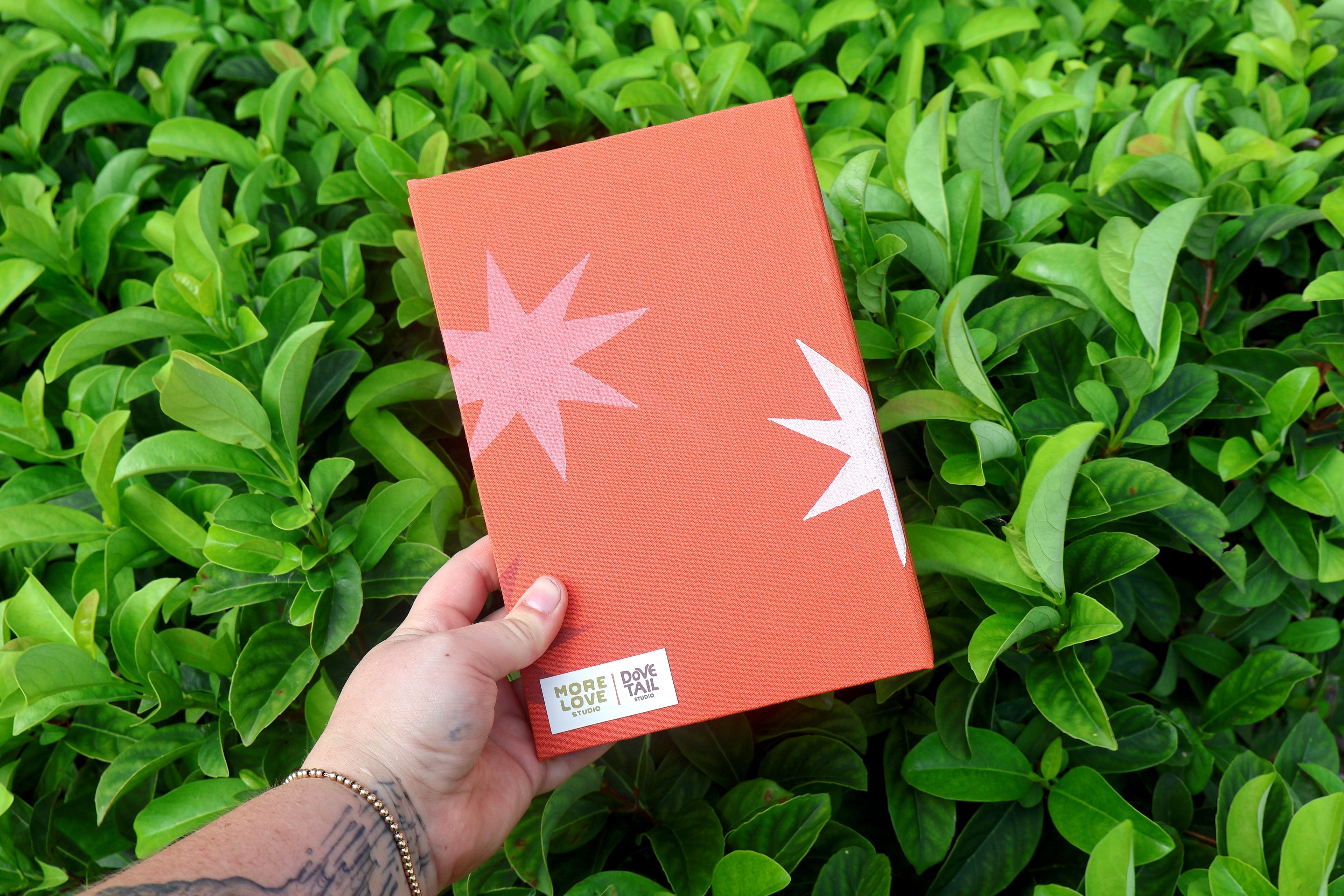 back cover of hand bound and linocut printed refillable journal with dovetail studio and more love studio branded tag. Orange cotton fabric cover with printed mauve and white starbursts.