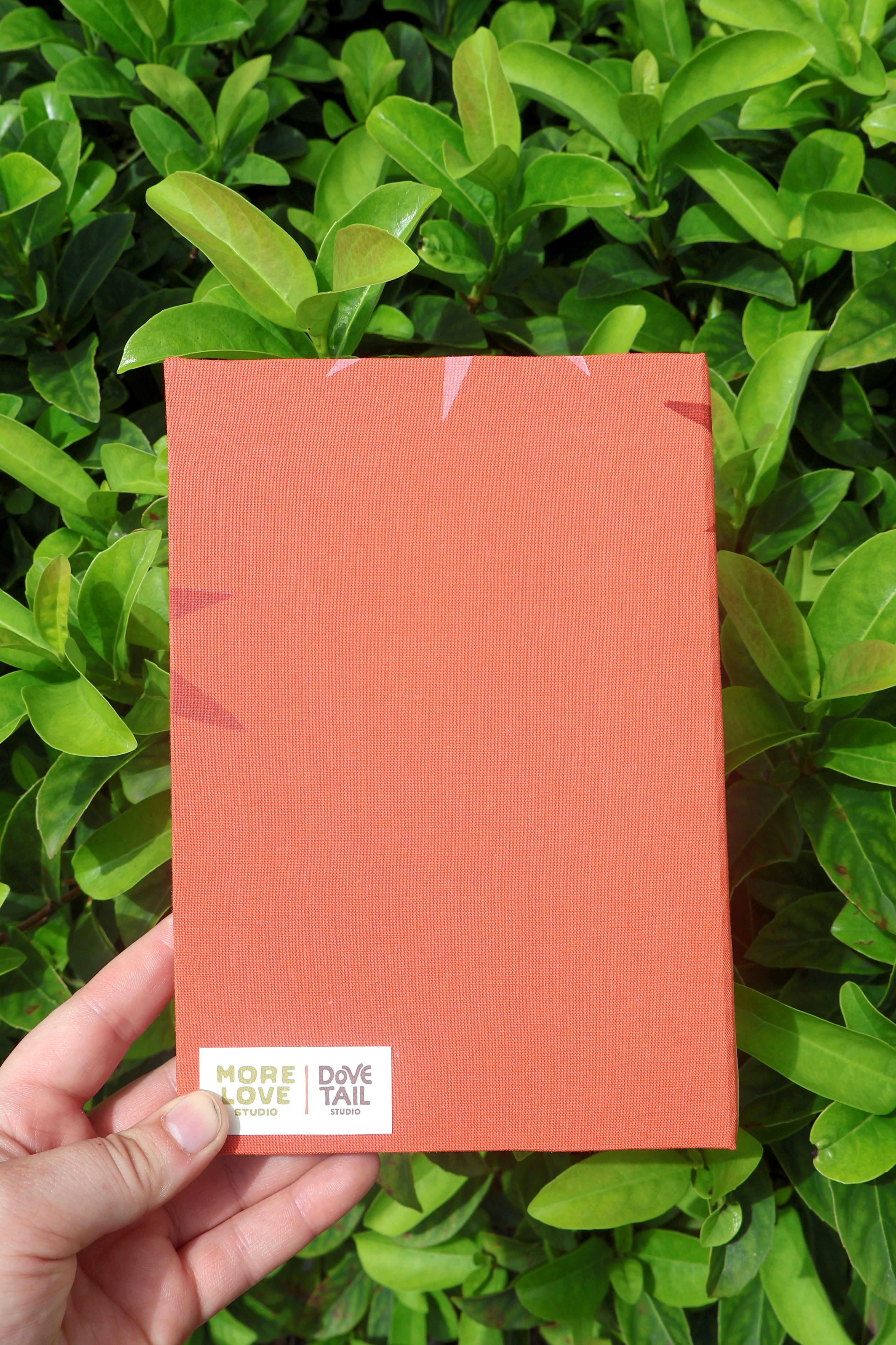 A hand holds a coral orange textured sketchbook against a background of green leaves. The bottom left corner features a white label with the text 'MORE LOVE STUDIO' and 'DOVE TAIL'.