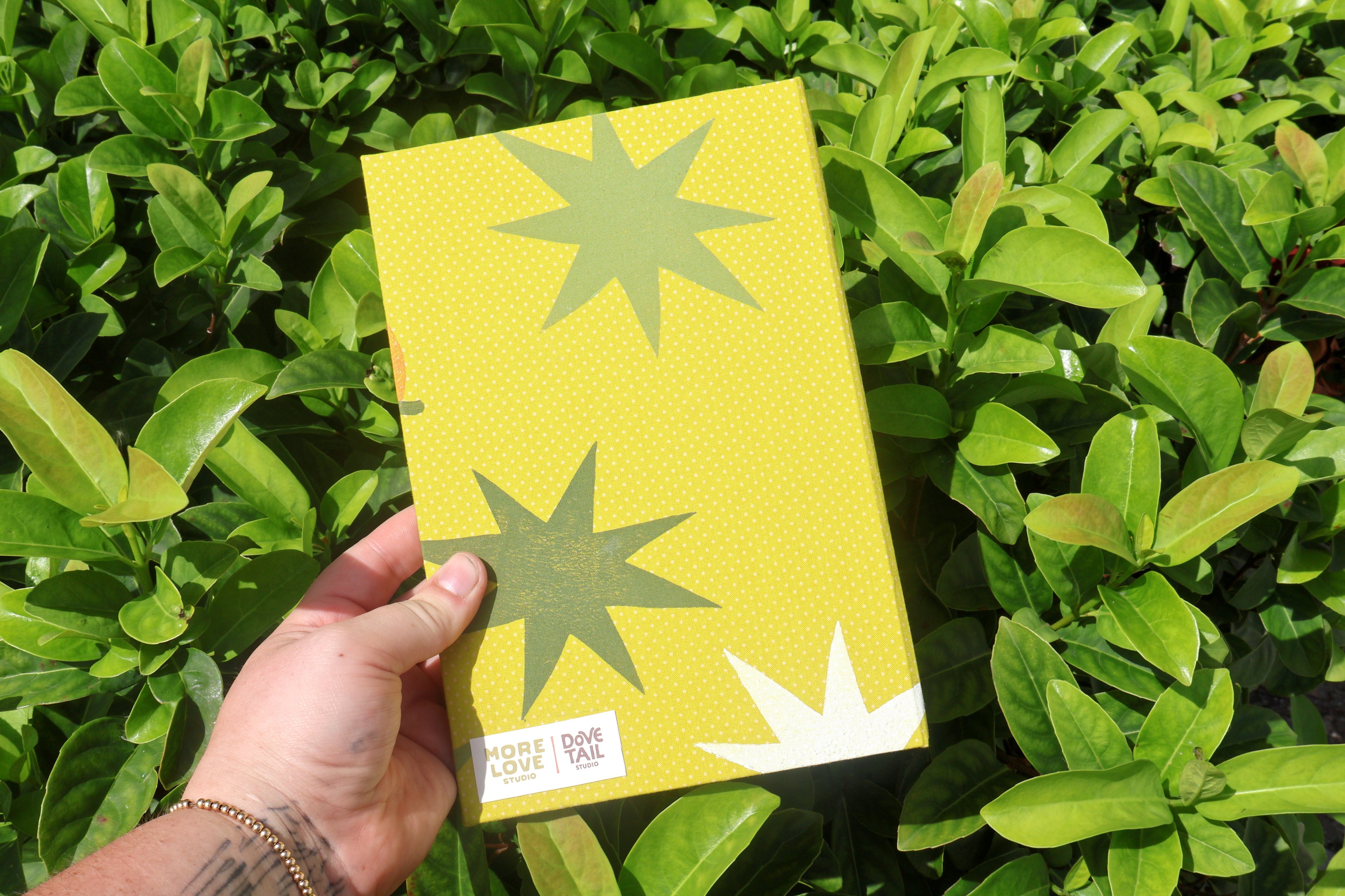 A hand holding a yellow textured notebook with green starburst patterns and a white label, set against a background of green foliage. hand printed and bound self care reflections log