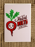 A square greeting card featuring a cartoon red beet with green leaves and the text 'My Heart BEETS FOR YOU' on a light pink background, photographed flat on a woven brown surface.