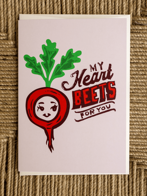 A square greeting card featuring a cartoon red beet with green leaves and the text 'My Heart BEETS FOR YOU' on a light pink background, photographed flat on a woven brown surface.