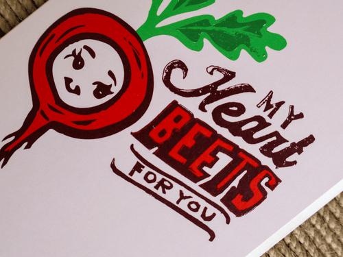 A close-up of a printed card or sign featuring a stylized red beet with green leaves and the phrase 'My Heart BEETS FOR YOU' in red and dark brown lettering on a white background.