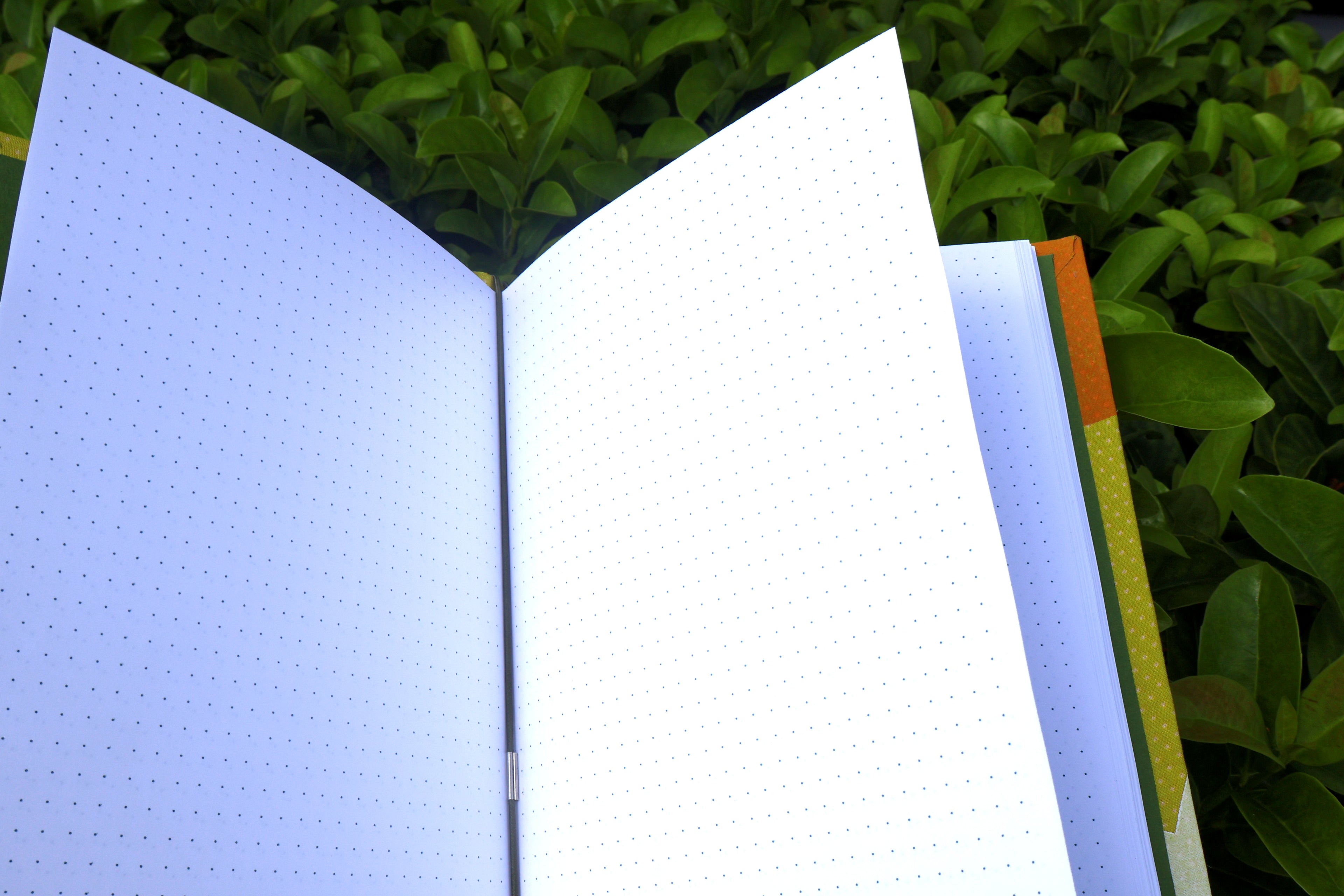 A hand holds opened yellow-patterned refillable bullet journals in front of dense green foliage. green interior cover paper and white bullet journal paper. hand bound and linocut printed with more love studio