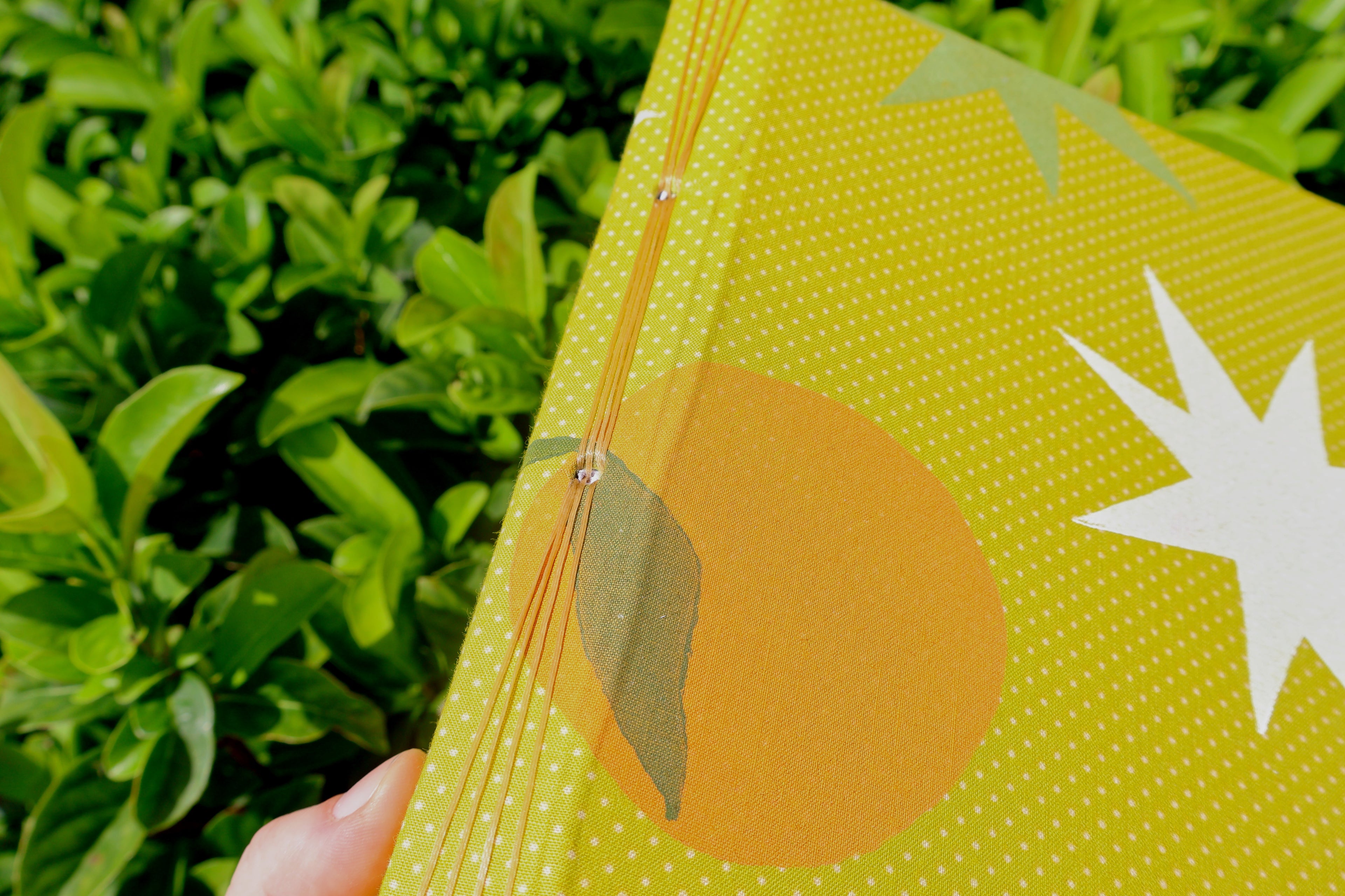 A close-up of a hand holding a yellow-green patterned sketchbook outdoors, with green foliage blurred in the background. The cover has a dotted pattern, a large orange circle, a white starburst, and a gray leaf motif. Hand-bound and printed with more love studio