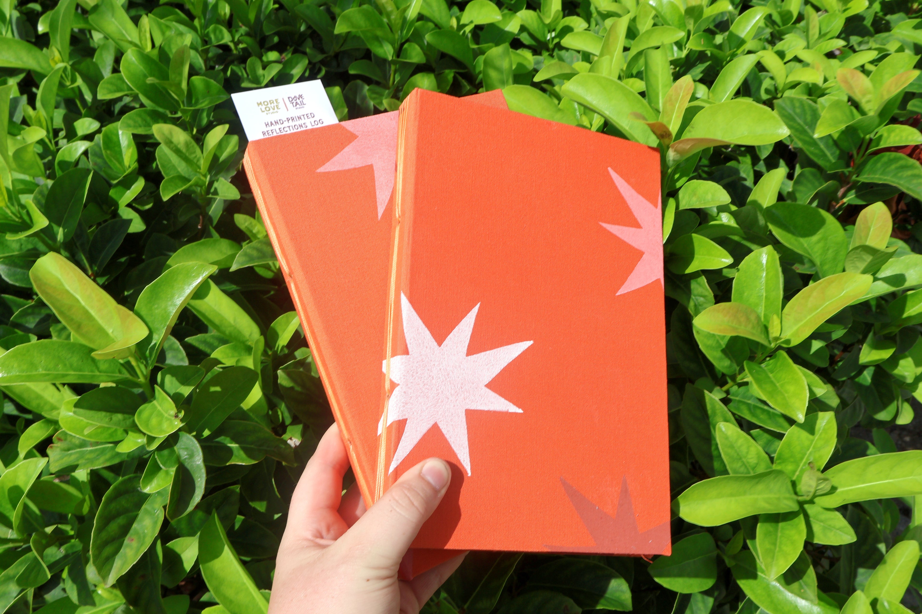 A hand holds two orange textured self care reflections logs decorated with orange and white starburst linocut prints, set against a background of green leaves. Hand printed linocut cover, hand bound self care reflections log