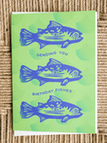 A square greeting card featuring three blue fish illustrations and the text 'SENDING YOU BIRTHDAY FISHES' on a bright green background, photographed on a woven beige surface.