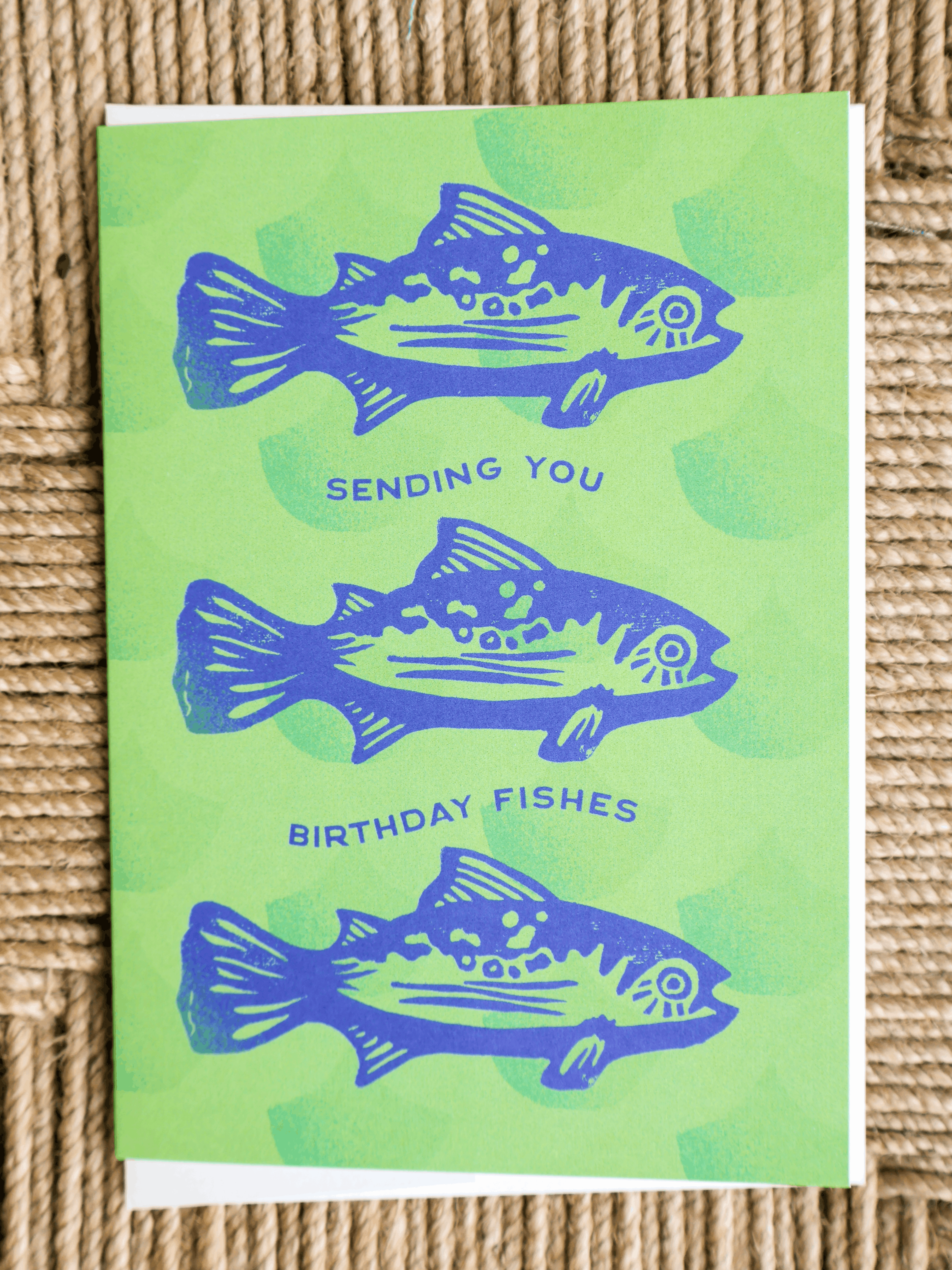 A square greeting card featuring three blue fish illustrations and the text 'SENDING YOU BIRTHDAY FISHES' on a bright green background, photographed on a woven beige surface.