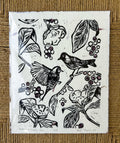 black birds on a branch with flowers surrounding them and purple blueberries on handmade cream paper handmade linocut print that has flower petals in it signed by artist on a textured brown background