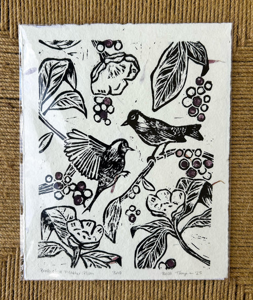black birds on a branch with flowers surrounding them and purple blueberries on handmade cream paper handmade linocut print that has flower petals in it signed by artist on a textured brown background