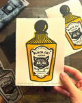 A hand holds a printed card featuring a stylized illustration of a black cat potion bottle in yellow, black, and white. Another matching print is visible in the background on a wooden surface, with art supplies partially visible around the edges.