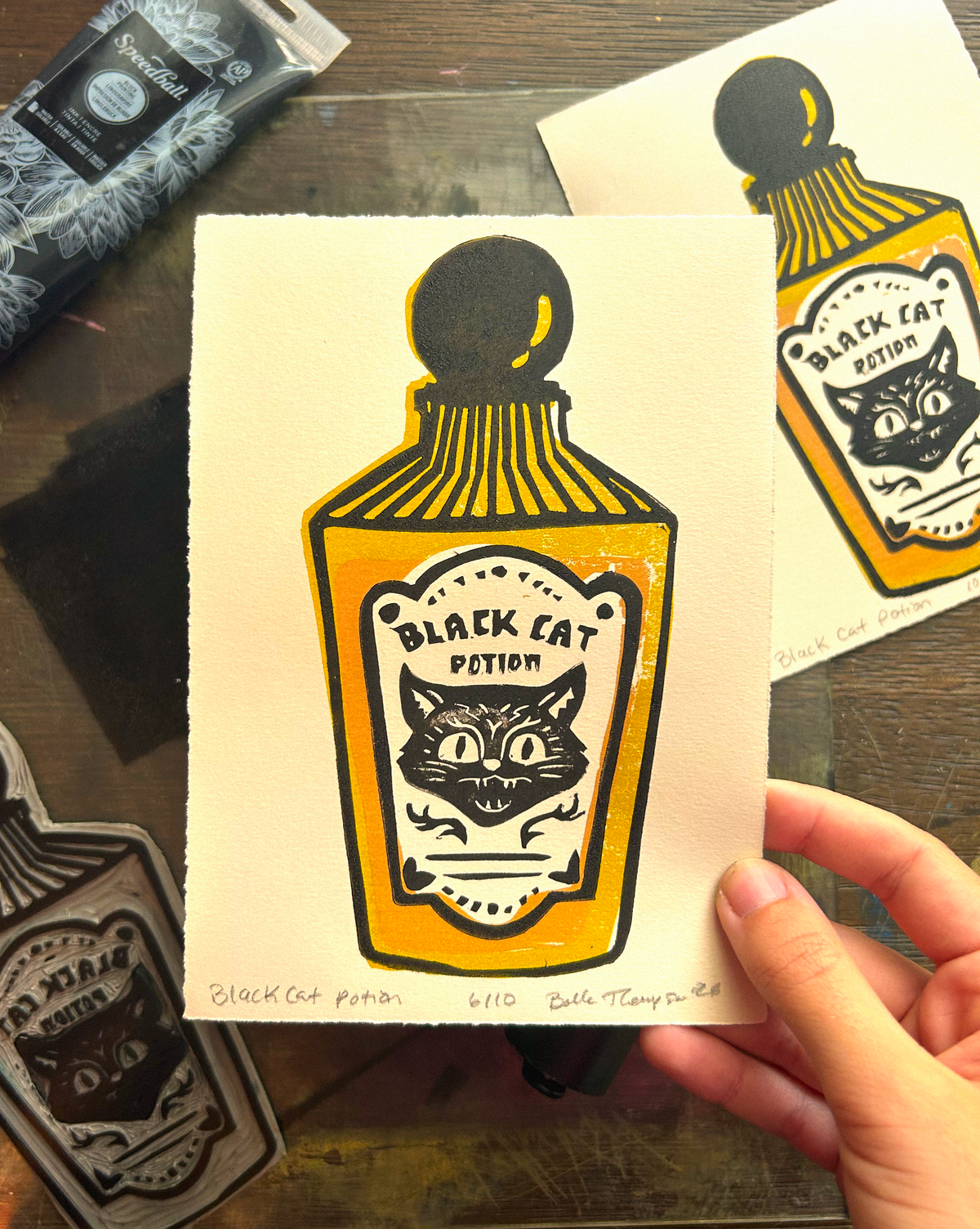 A hand holds a printed card featuring a stylized illustration of a black cat potion bottle in yellow, black, and white. Another matching print is visible in the background on a wooden surface, with art supplies partially visible around the edges.