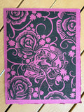 decorative black skull with black rose flowers growing out of it on pink paper using negative space handmade linocut print on heavyweight paper signed by artist with stripped wooden 