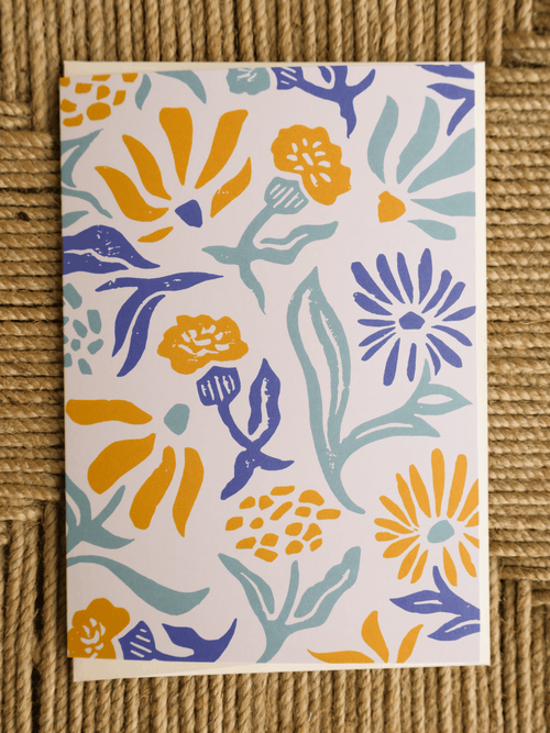 A rectangular art print or card featuring a colorful floral pattern, displayed flat on a textured brown woven mat. The pattern contains yellow, blue, and light teal flowers and leaves on a white background.