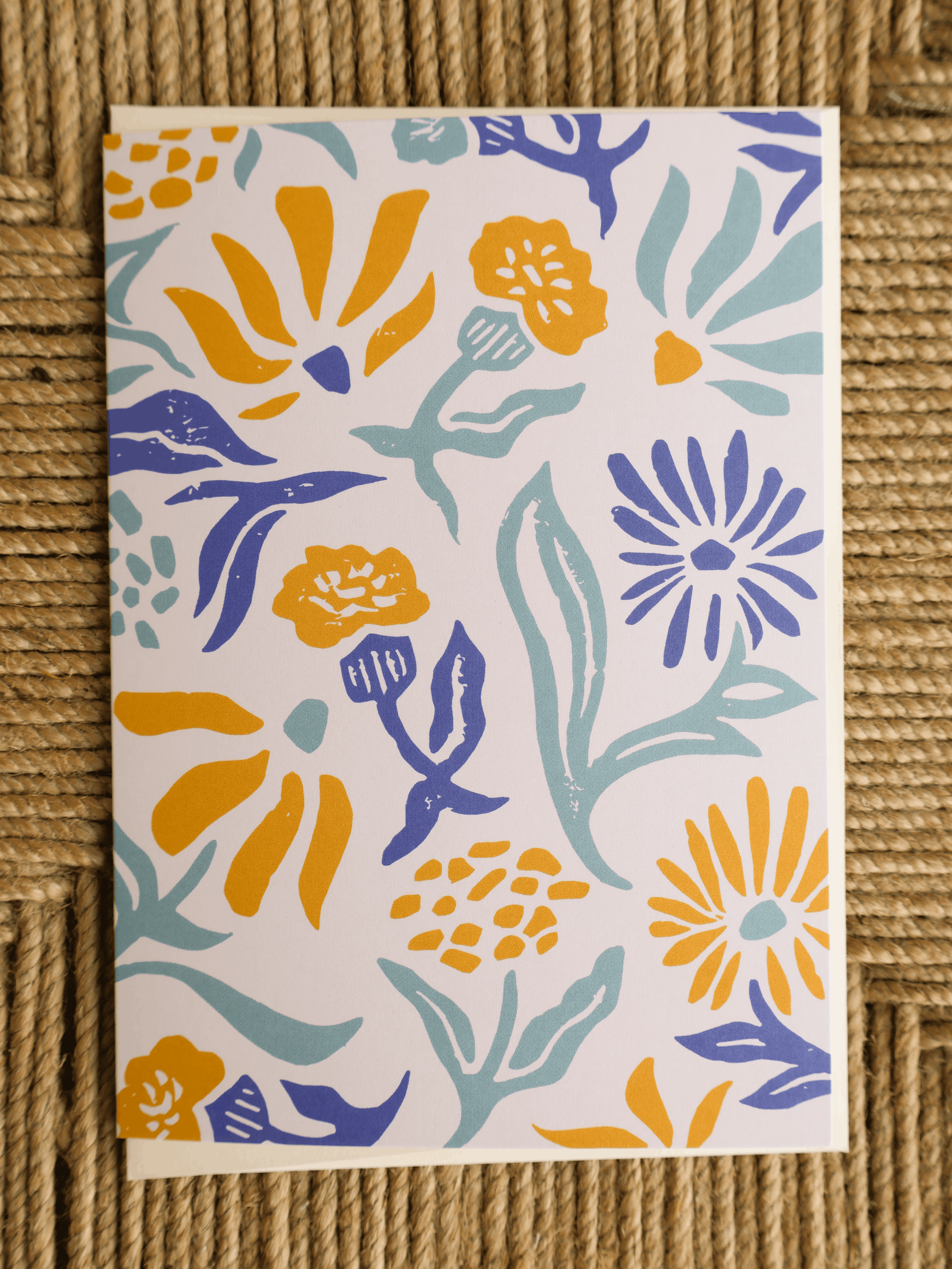 A rectangular art print or card featuring a colorful floral pattern, displayed flat on a textured brown woven mat. The pattern contains yellow, blue, and light teal flowers and leaves on a white background.