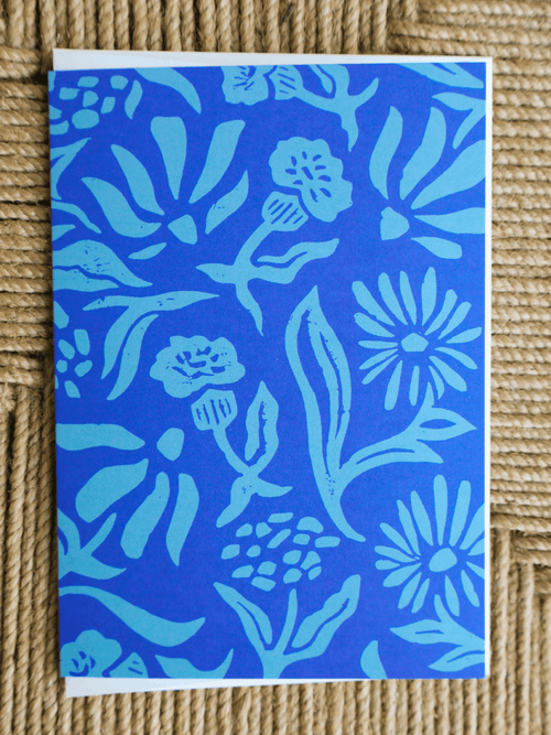 A square art print or card featuring a bold floral pattern in shades of blue and light cyan, photographed flat on a woven beige surface.
