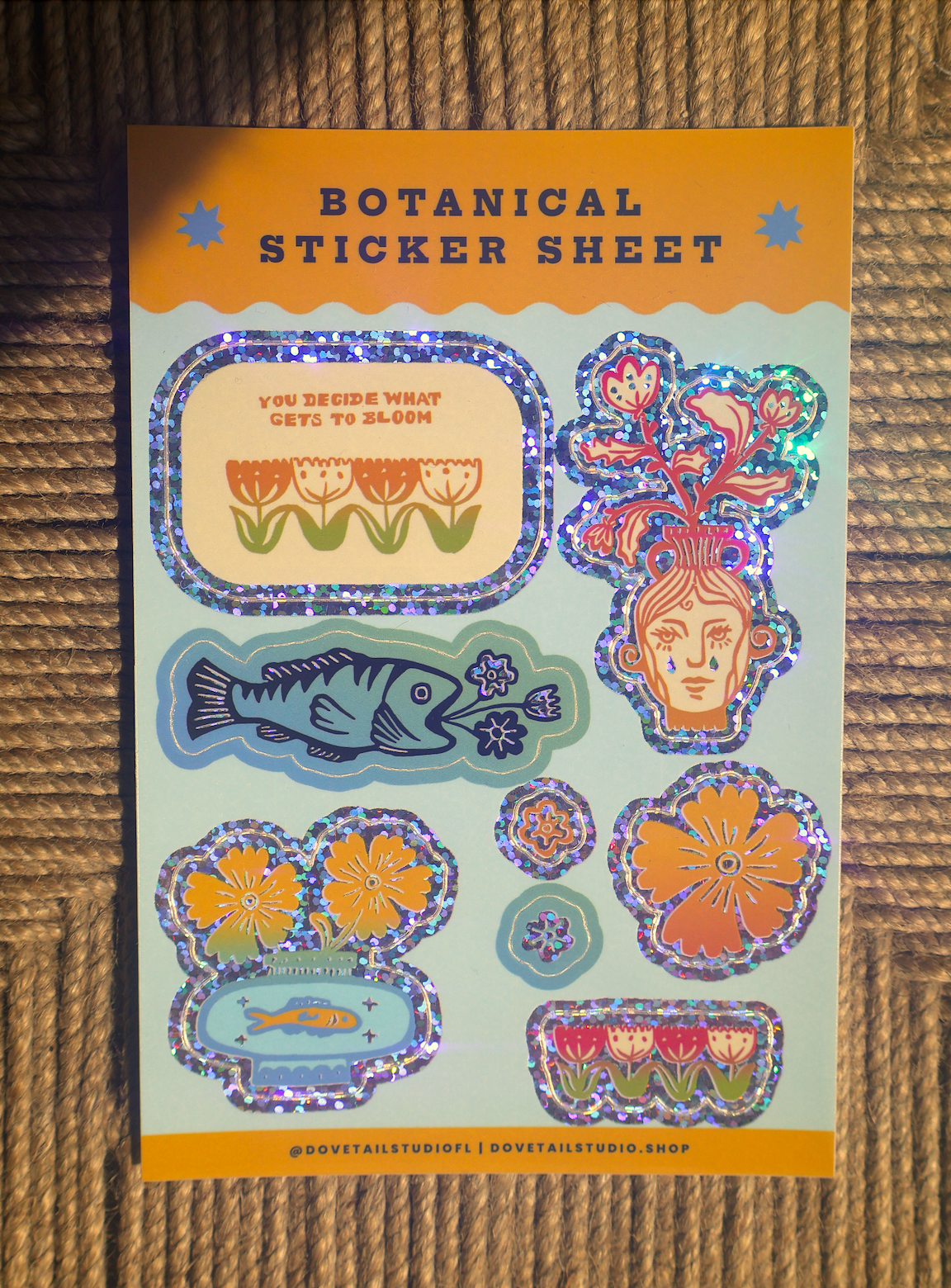 A botanical sticker sheet featuring whimsical illustrated stickers including flowers, fish, and a face with coral-like hair, printed on a pastel card with an orange header that reads 'BOTANICAL STICKER SHEET'. The sheet is photographed flat on a woven brown surface.