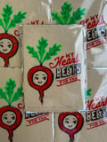 handmade linocut prints showing beets with faces that say my heart beets for you in the colors red, maroon, and green on tan paper signed by artist layered in the background