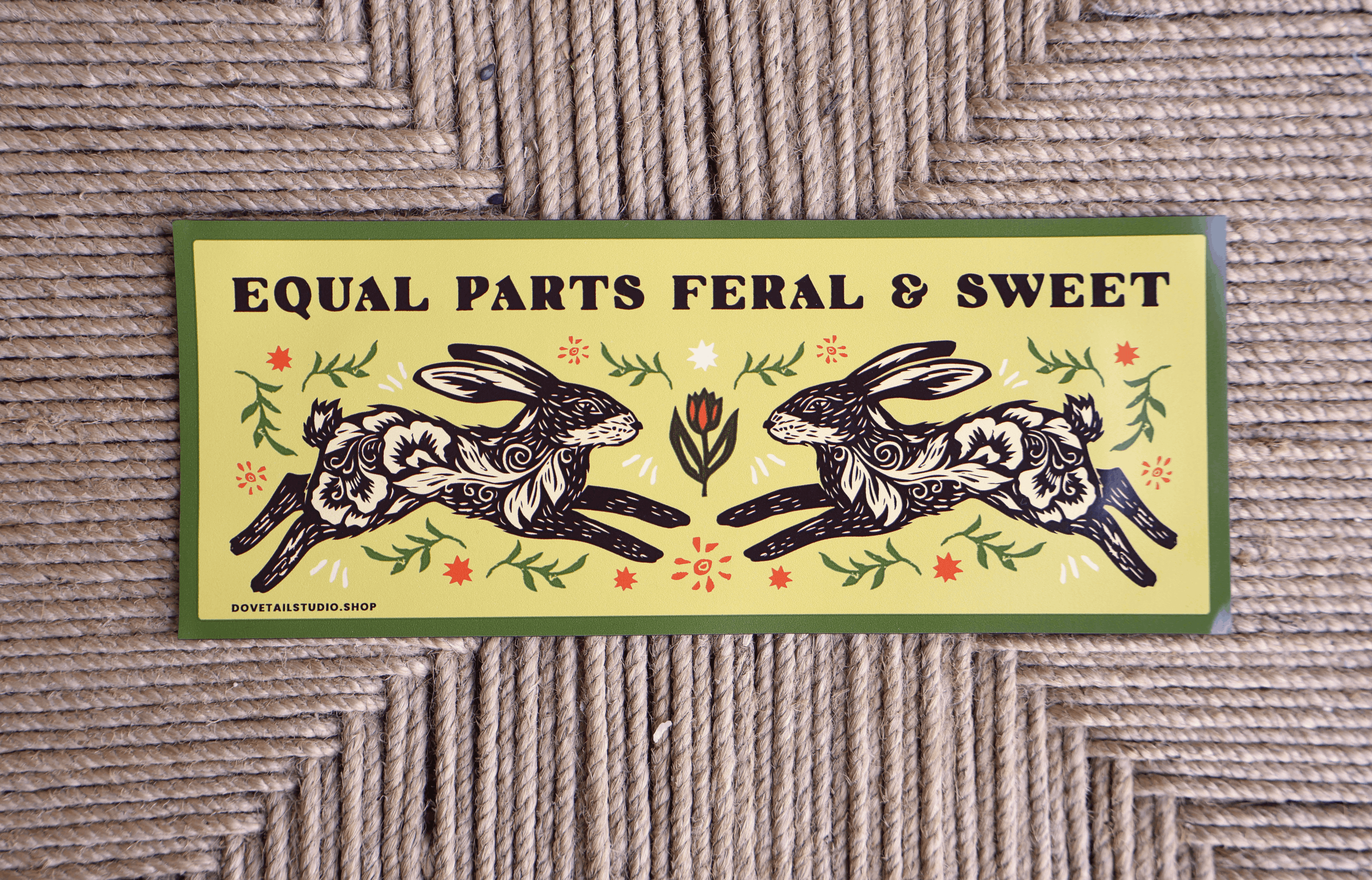 A rectangular decorative sign or print with the text 'EQUAL PARTS FERAL & SWEET' above an illustration of two rabbits facing each other, surrounded by floral and leaf motifs. The sign has a green border and is photographed on a woven rope surface.