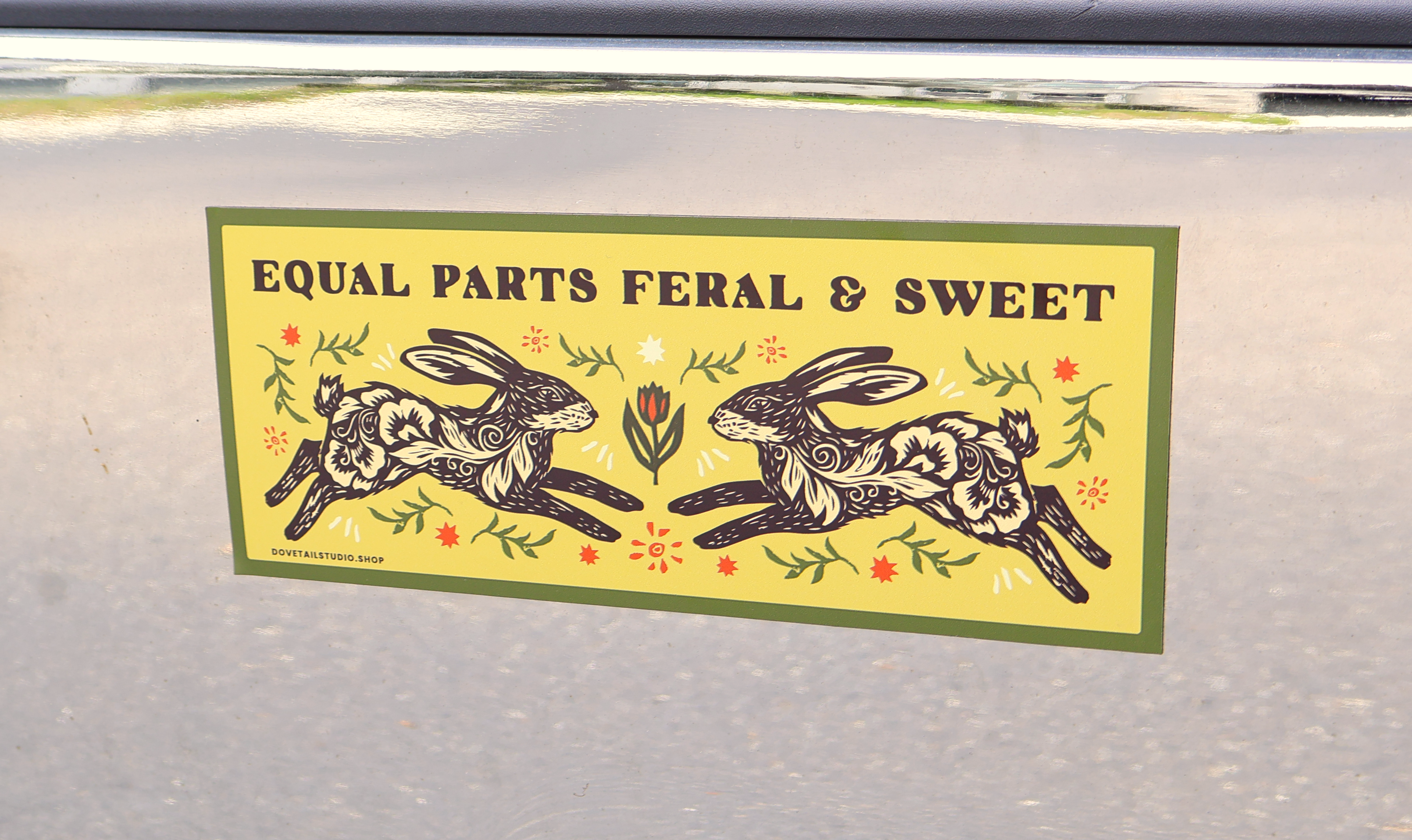 A rectangular sticker with the text 'EQUAL PARTS FERAL & SWEET' and an illustrated design of two rabbits facing each other, placed on a gray surface.