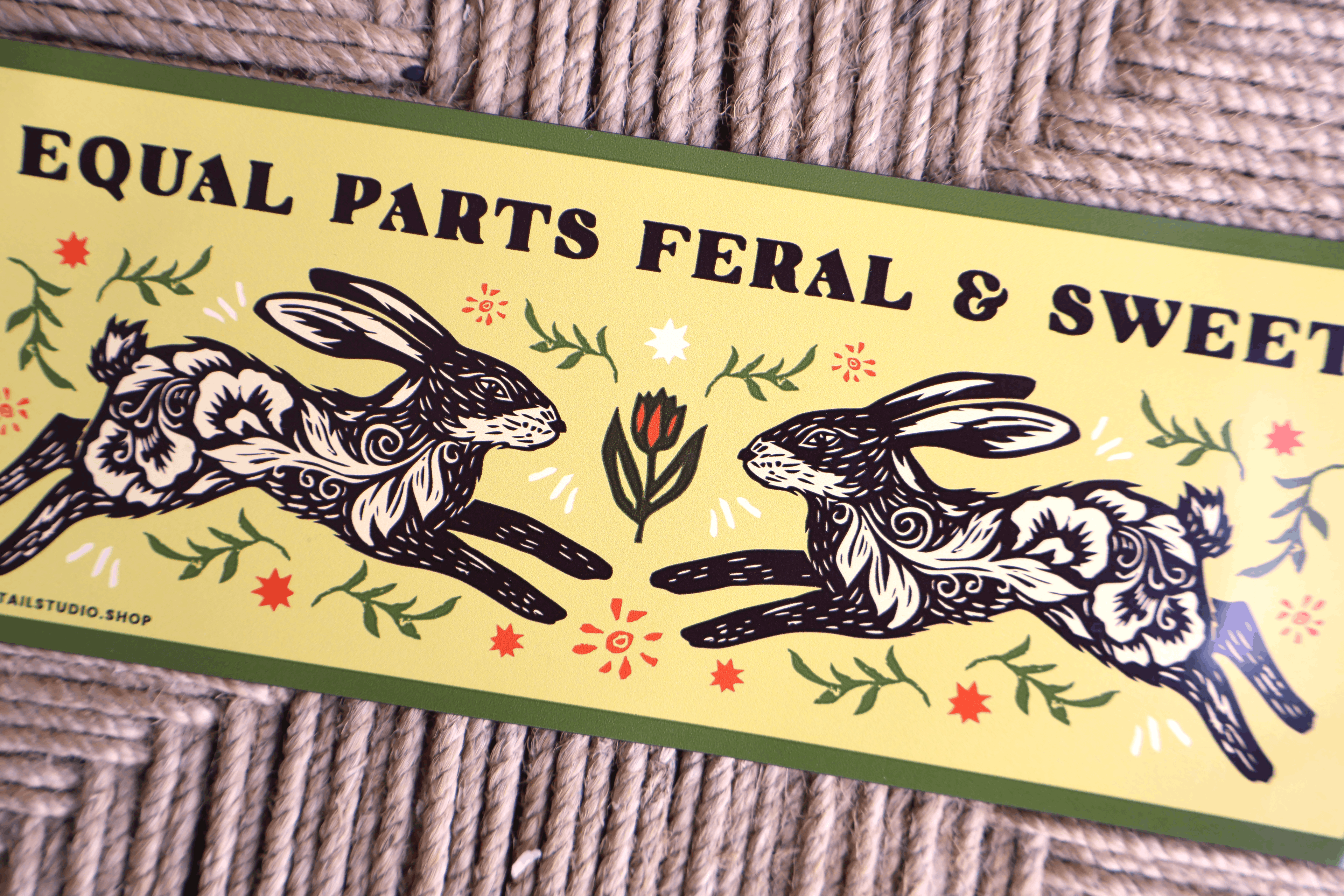 A rectangular printed label or sign with the text 'EQUAL PARTS FERAL & SWEET' above two stylized rabbits facing each other, placed on a woven rope surface. The design includes floral motifs and green border lines.