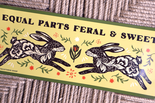 A rectangular printed label or sign with the text 'EQUAL PARTS FERAL & SWEET' above two stylized rabbits facing each other, placed on a woven rope surface. The design includes floral motifs and green border lines.