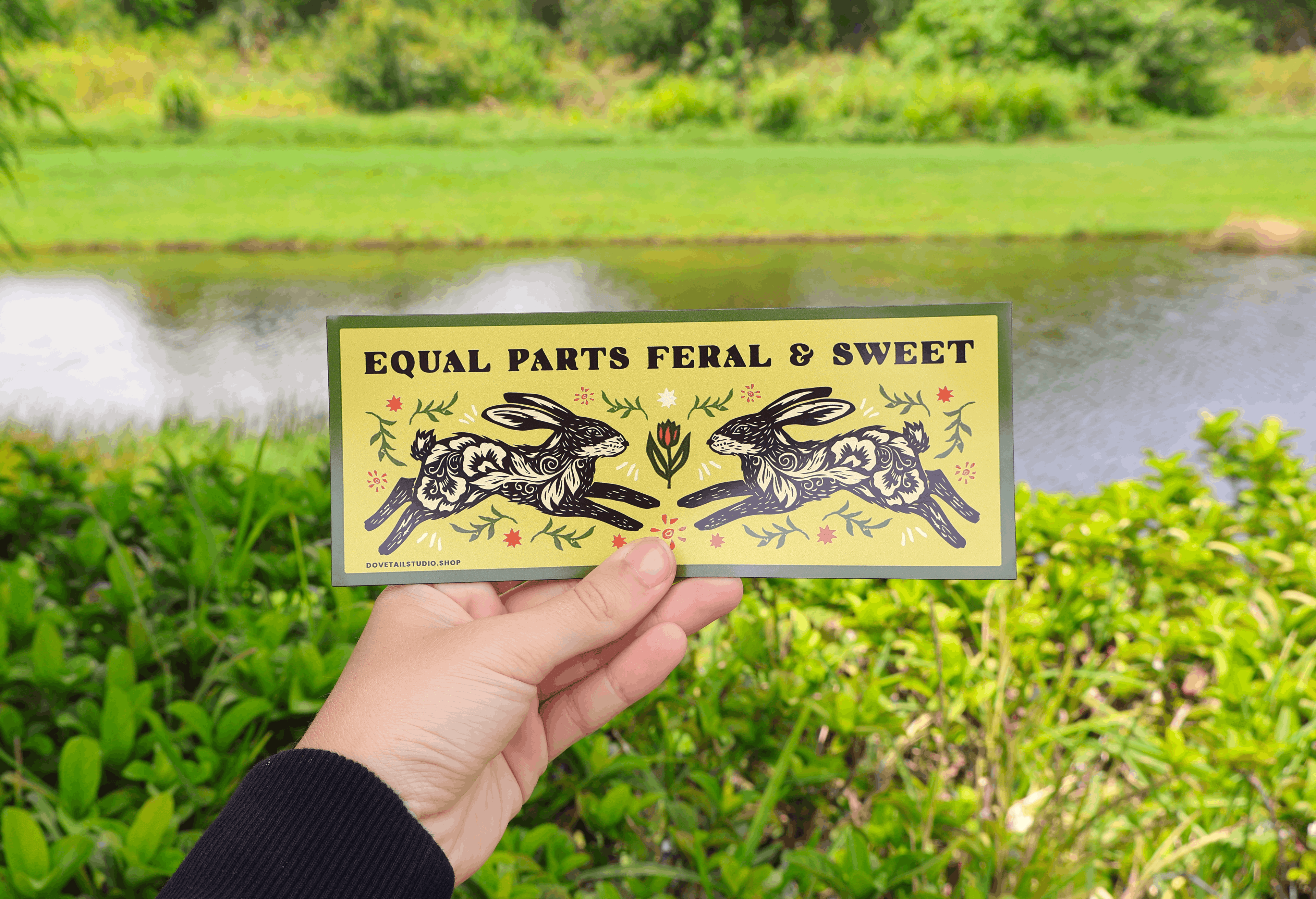 A hand holds a rectangular art print featuring two black-and-white rabbits and the text 'EQUAL PARTS FERAL & SWEET' against a blurred outdoor background of green foliage and a pond.