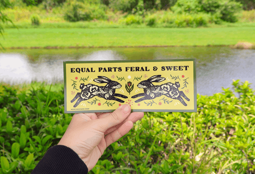A hand holds a rectangular art print featuring two black-and-white rabbits and the text 'EQUAL PARTS FERAL & SWEET' against a blurred outdoor background of green foliage and a pond.