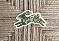 A die-cut sticker featuring a stylized green rabbit illustration with floral and swirl patterns on a cream background, placed on a woven jute surface.