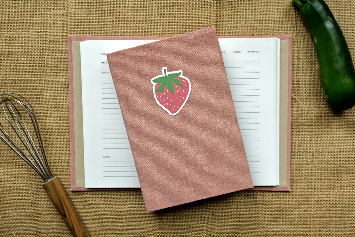 A pink textured hardcover recipe log with a die-cut strawberry linocut design on the cover, placed on top of an open recipe log journal. The scene is styled as a flat lay on a burlap surface with a whisk on the left and a green vegetable on the right.
