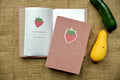 A pink textured hardcover recipe log with a strawberry linocut print design on the cover, shown next to an open matching book on a burlap background with a green cucumber and a yellow squash as props.