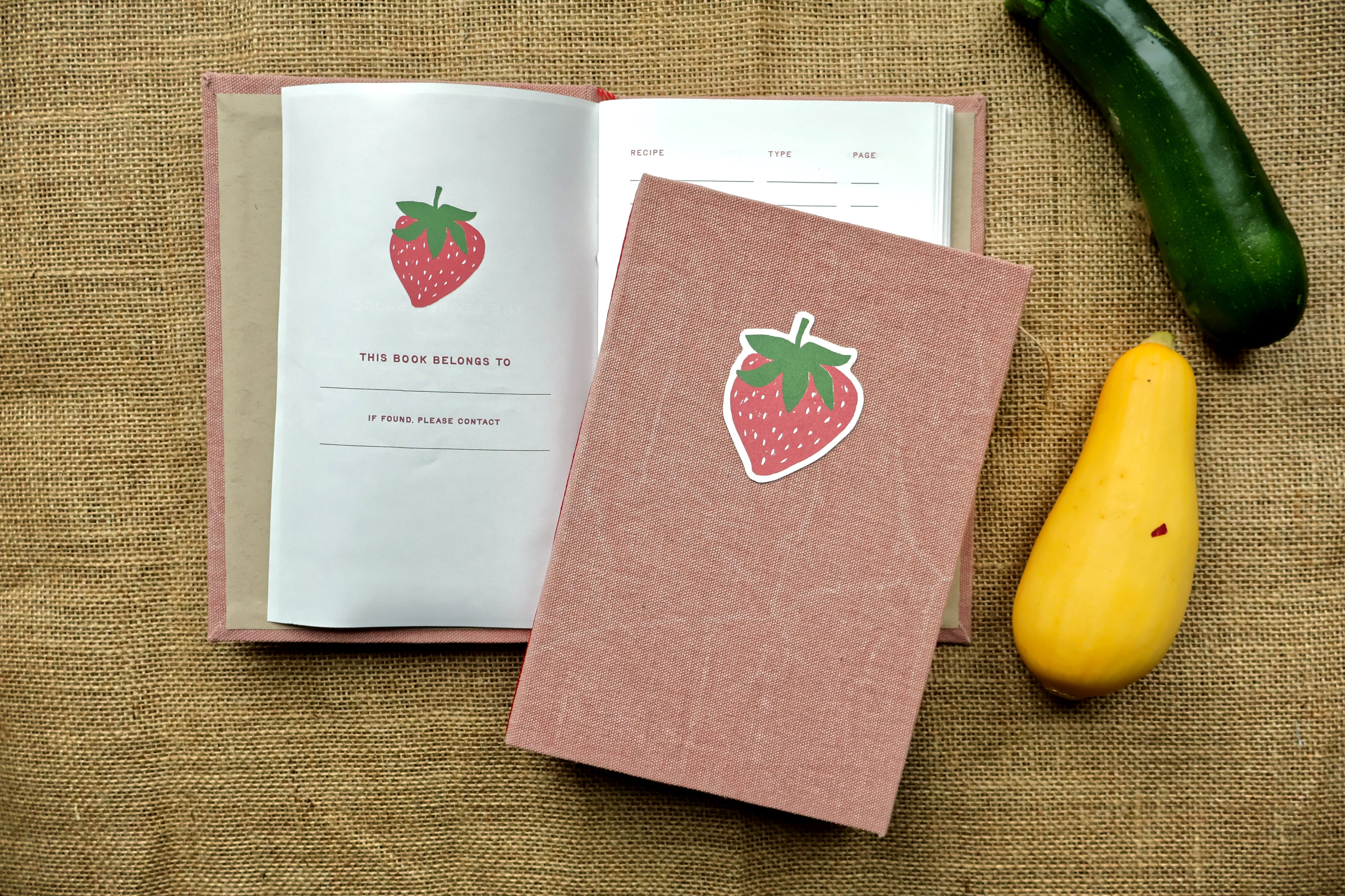 A pink textured hardcover recipe log with a strawberry linocut print design on the cover, shown next to an open matching book on a burlap background with a green cucumber and a yellow squash as props.