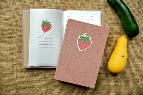 A pink textured hardcover recipe log with a strawberry linocut print design on the cover, shown next to an open matching book on a burlap background with a green cucumber and a yellow squash as props.