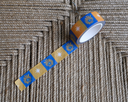 linocut design of checkered blue, gold, and cream organic flowers and star on decorative washi tape. unrolled showing the design on a textured brown background. by dovetail studio
