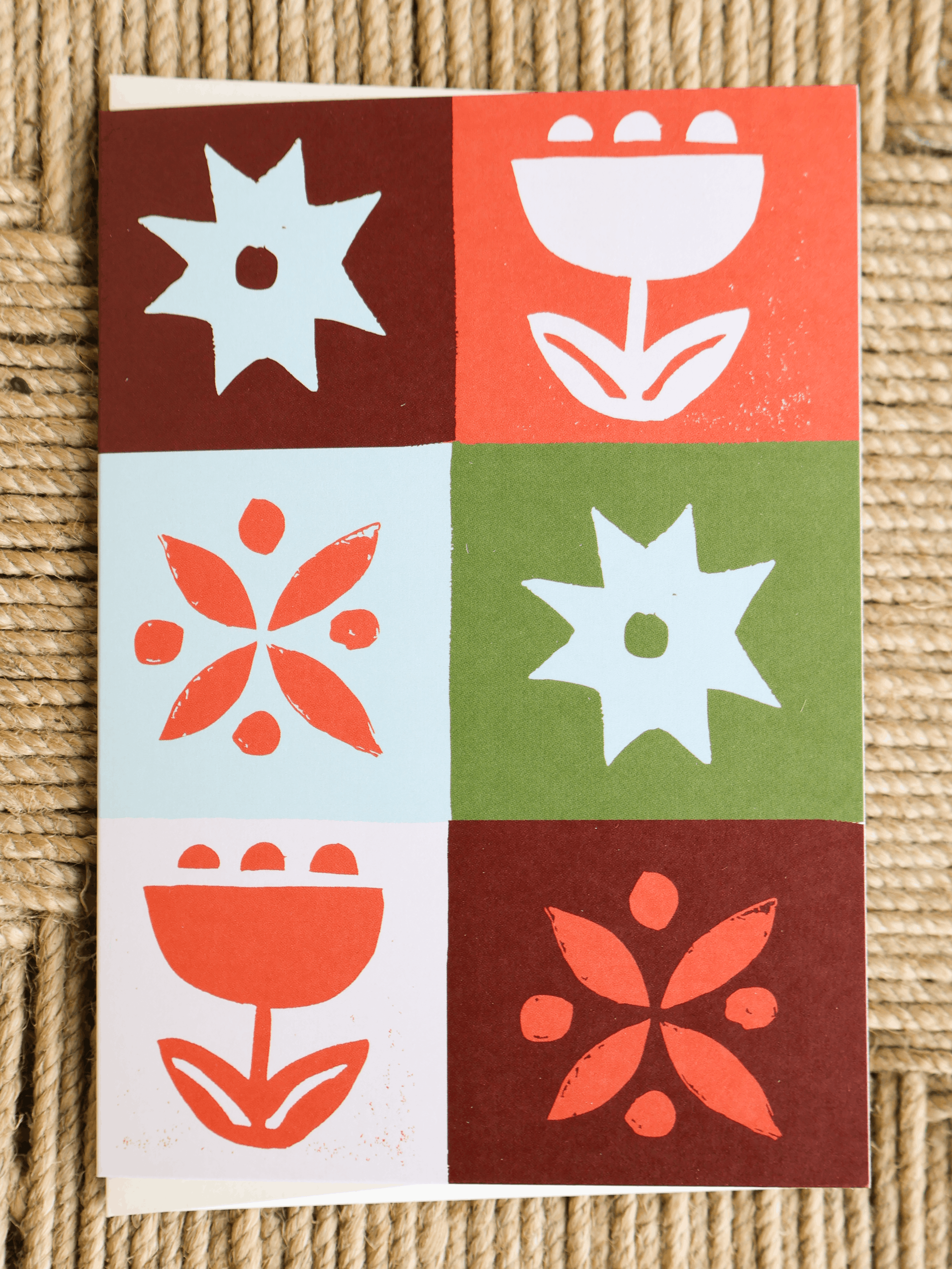 A square art print or card featuring a 2x3 grid of stylized floral and starburst motifs in coral, white, green, and dark red, photographed on a woven beige surface.