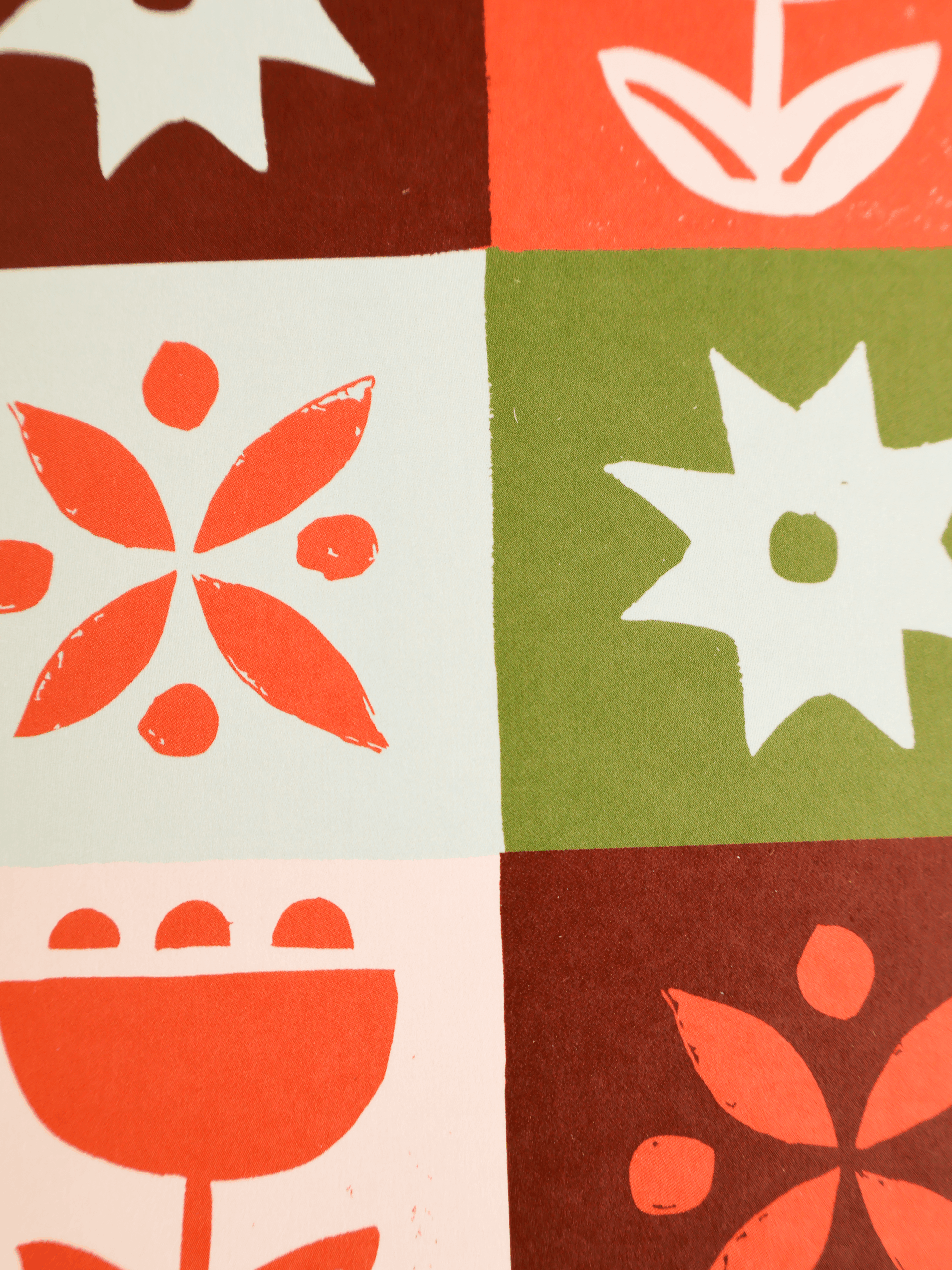 A close-up of a textile or wallpaper pattern featuring a 2x3 grid of blocks with retro floral and geometric linocut-style motifs in coral orange, olive green, dark brown, and off-white.