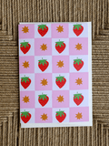 A square card featuring a pink and white checkerboard pattern with red strawberries, green leafy tops, and orange starbursts, photographed on a woven brown surface.