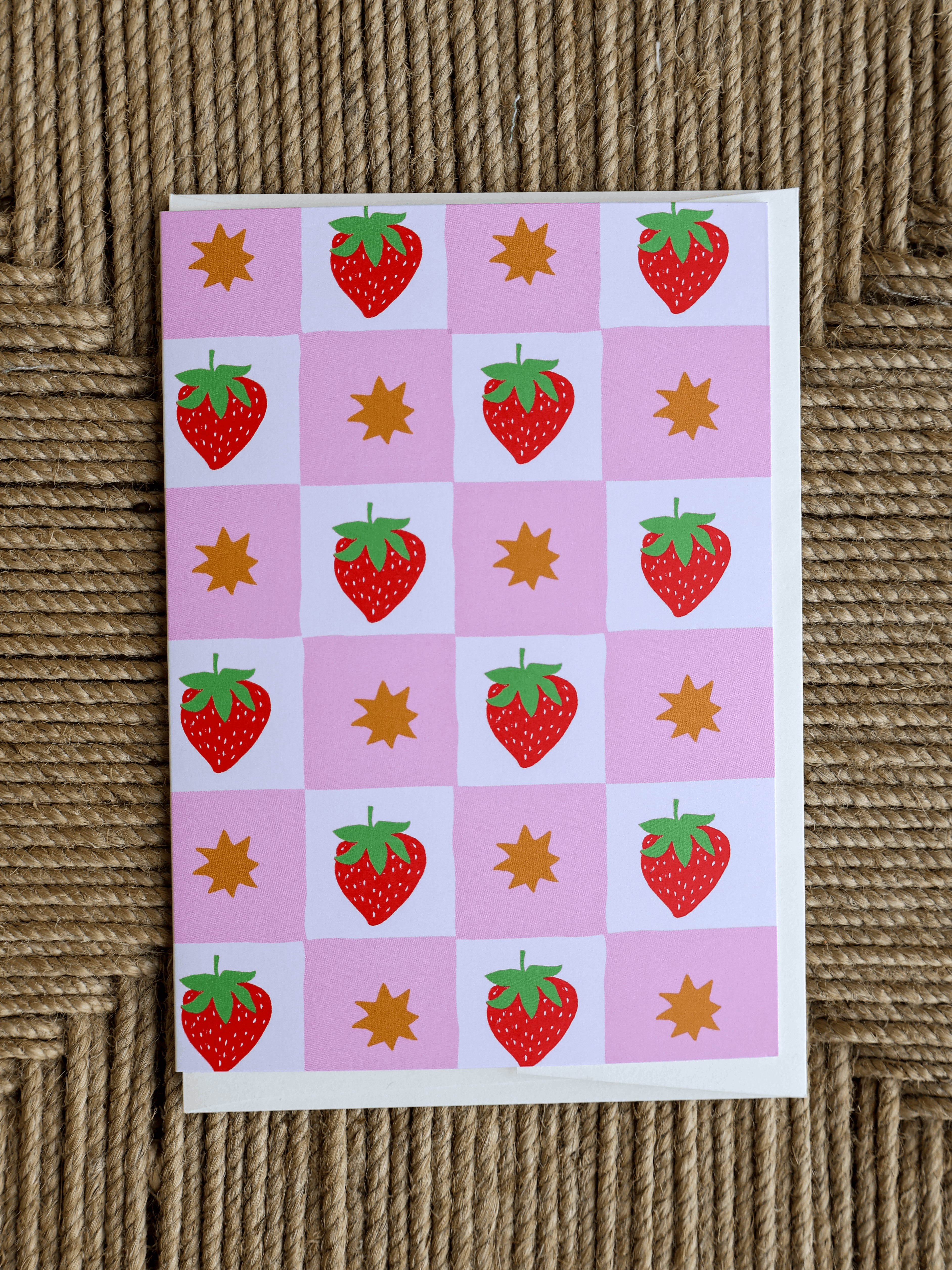 A square card featuring a pink and white checkerboard pattern with red strawberries, green leafy tops, and orange starbursts, photographed on a woven brown surface.