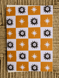 A square card or print featuring a checkerboard pattern of orange and white squares, each containing a black sun face or white starburst motif, photographed on a woven beige surface.