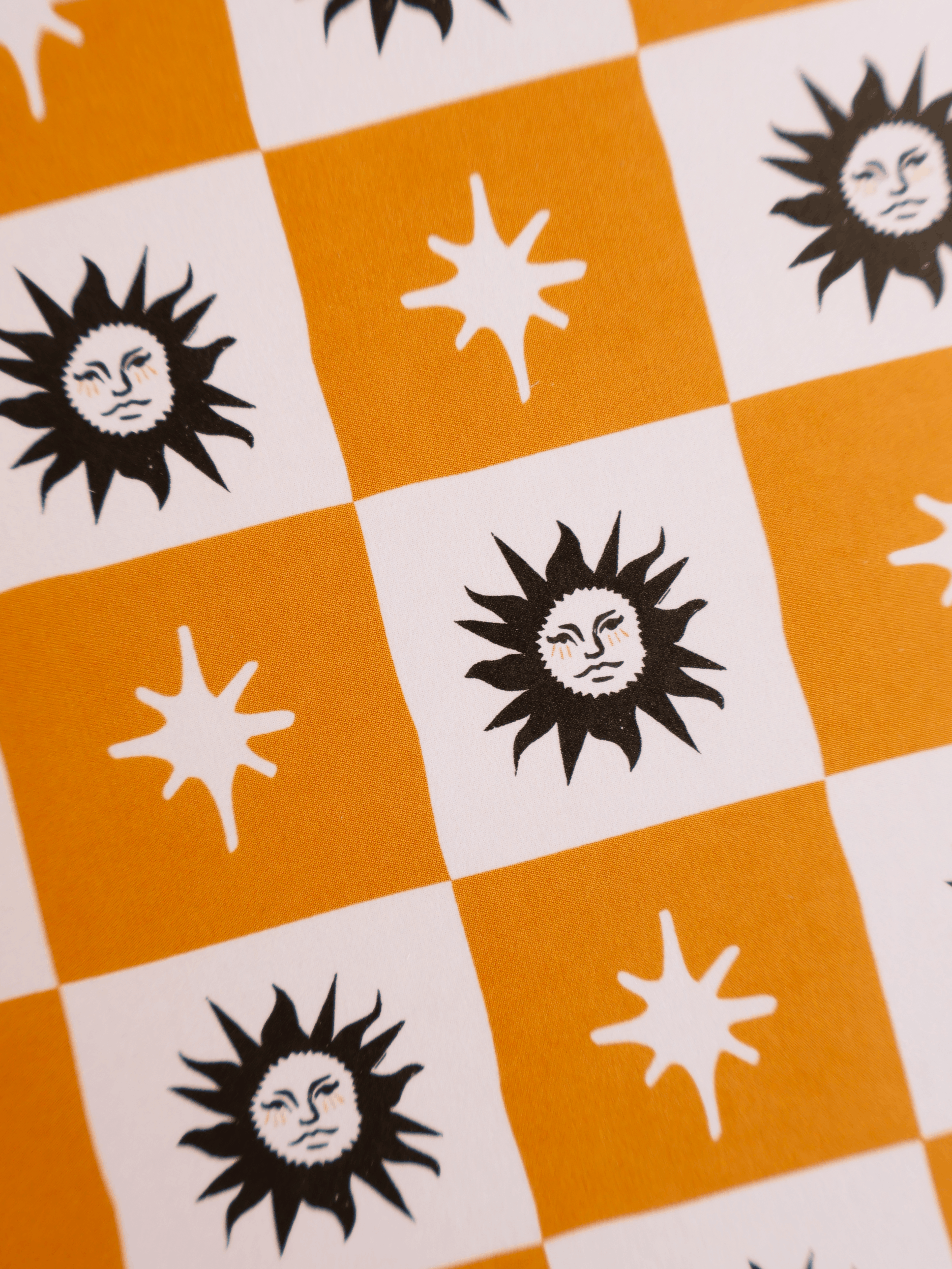 A close-up of a patterned fabric or wallpaper featuring alternating orange and white checkered squares with black sun faces and white starburst motifs.