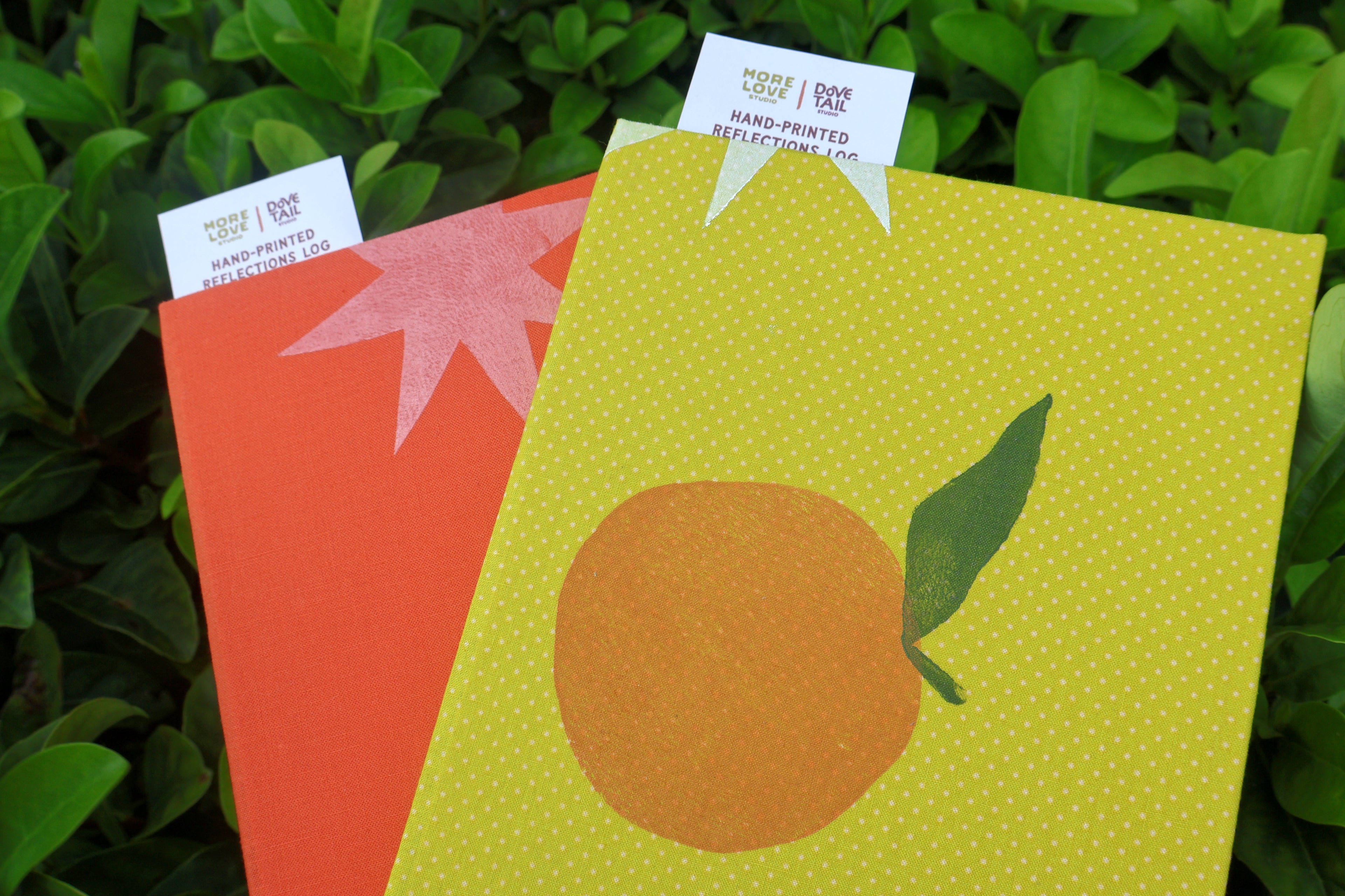 Two hand-printed self care reflection logs displayed outdoors against green foliage. The front item is yellow with a printed orange and leaf, and the back visible item is orange with a pink starburst. hand bound and linocut printed with more love studio